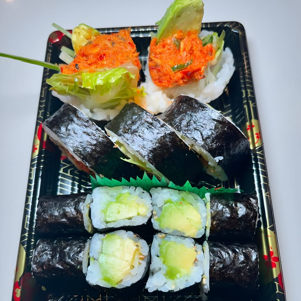 A black tray of Sushi Guys sushi, including spicy tuna, avocado rolls, and nori-wrapped rolls.