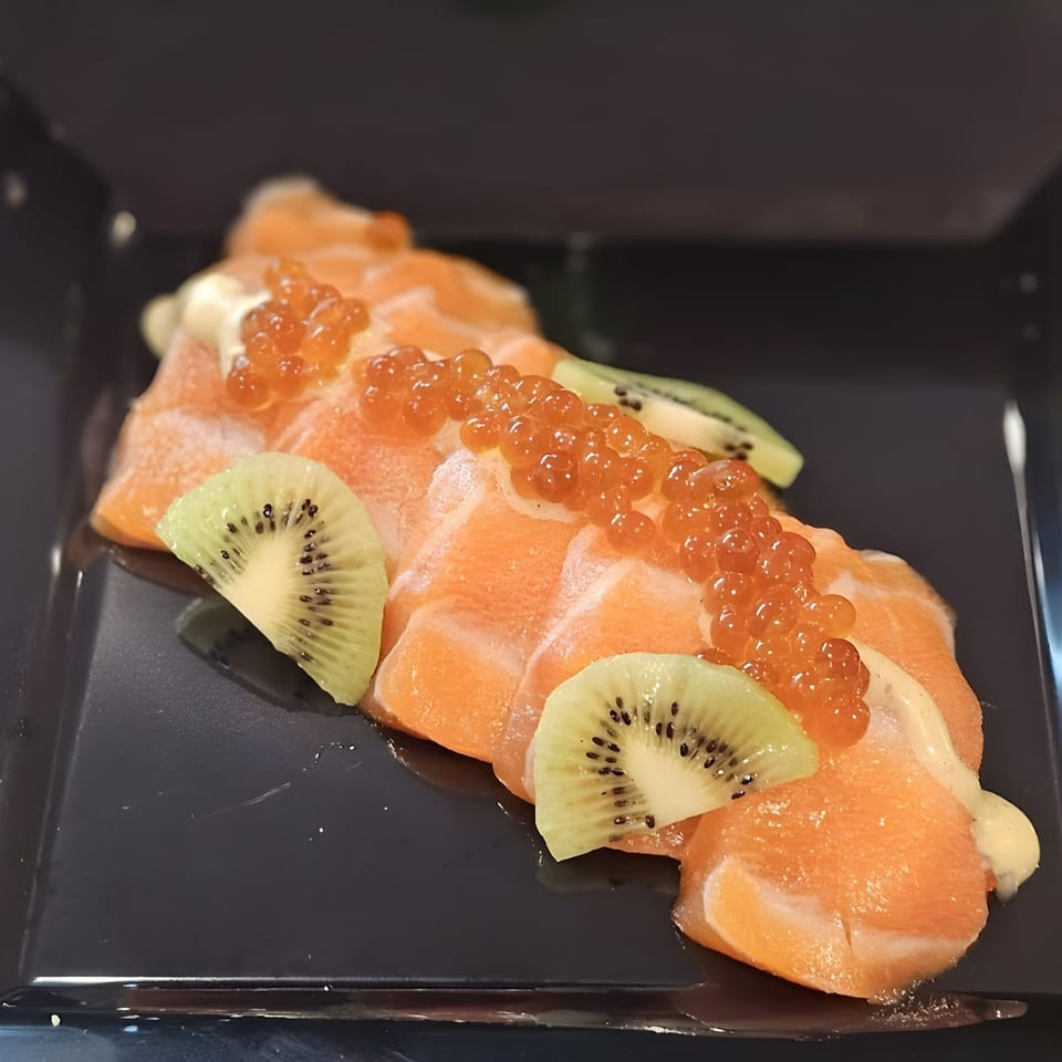 Salmon sashimi topped with bright orange roe, white sauce, and kiwi slices on a dark plate.